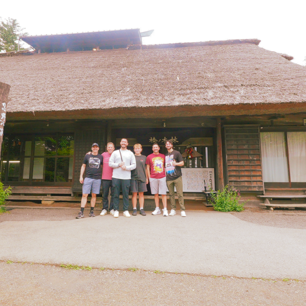 Six American team members standing in front of a traditional folk house in Oshino Hakkai during a photo tour near Mt. Fuji.